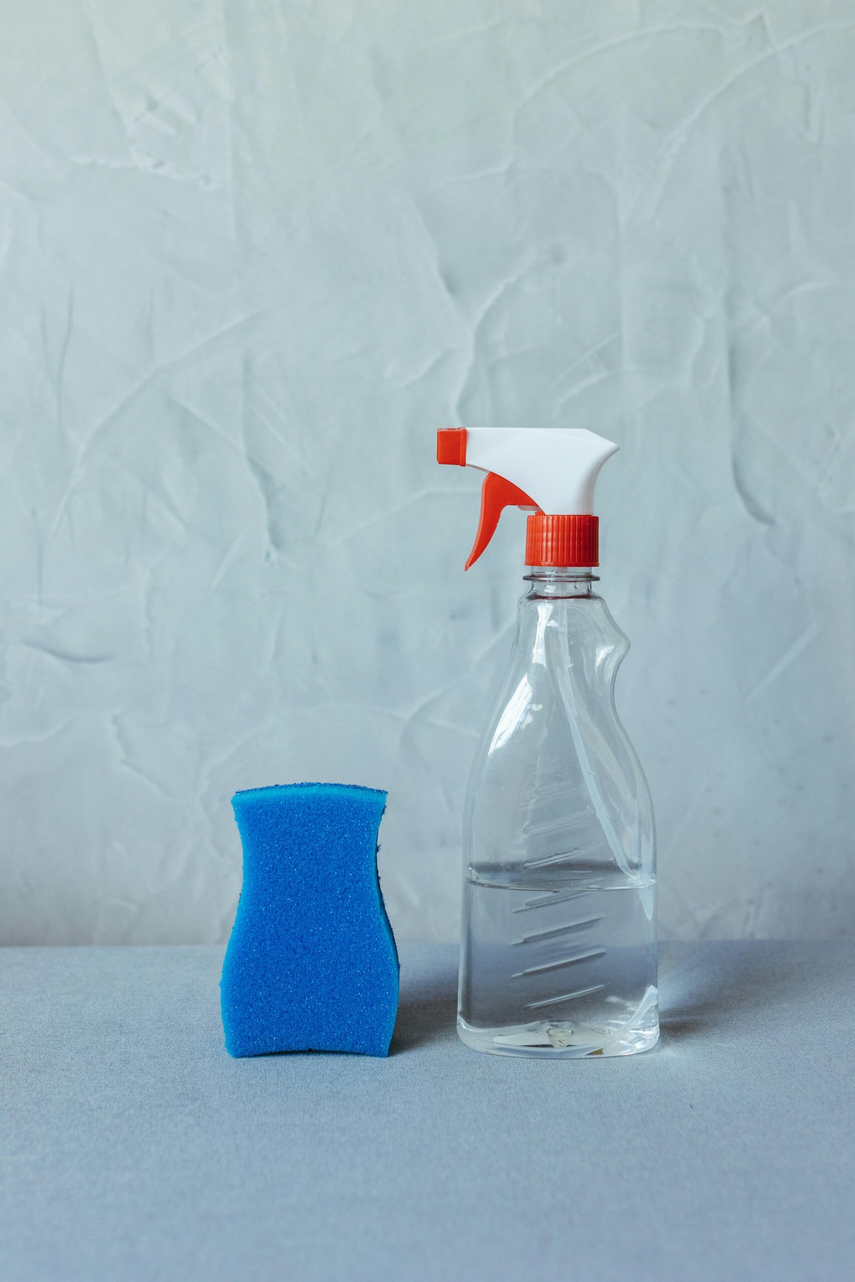A clear spray bottle and blue sponge on a textured surface for cleaning tasks.